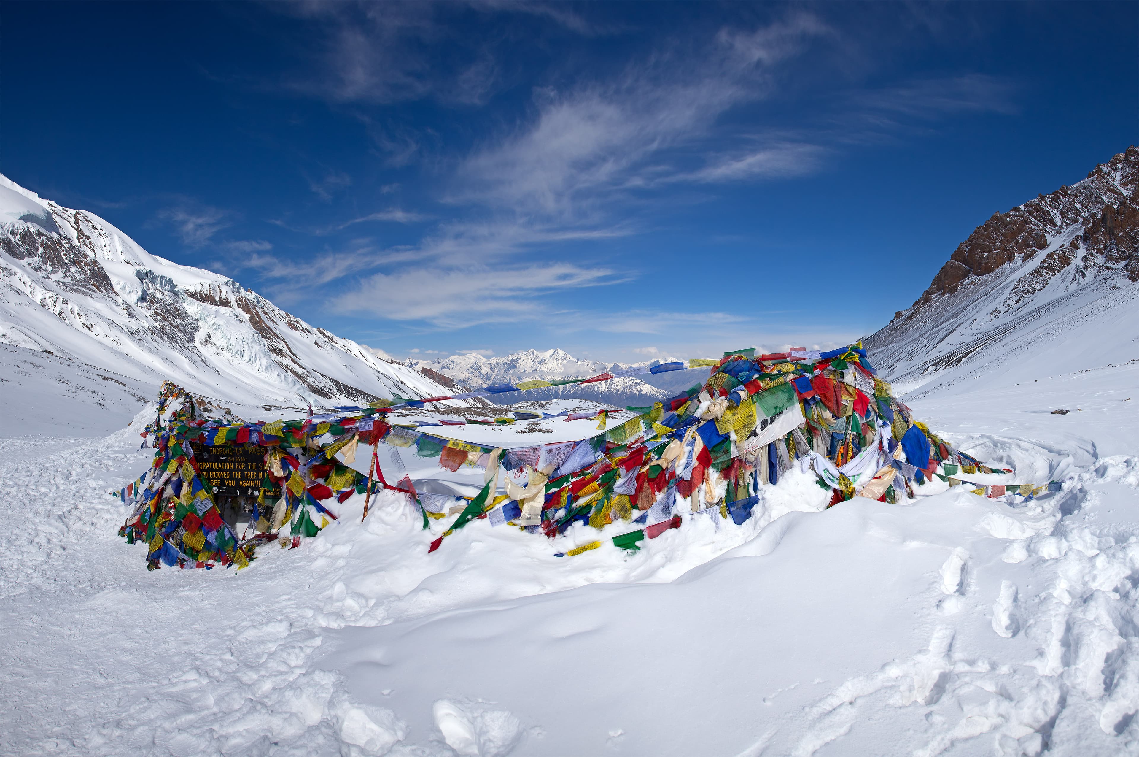 Tibetan prayer flags flapping in alpine winds at the summit of Thorong La Pass with towering snow peaks